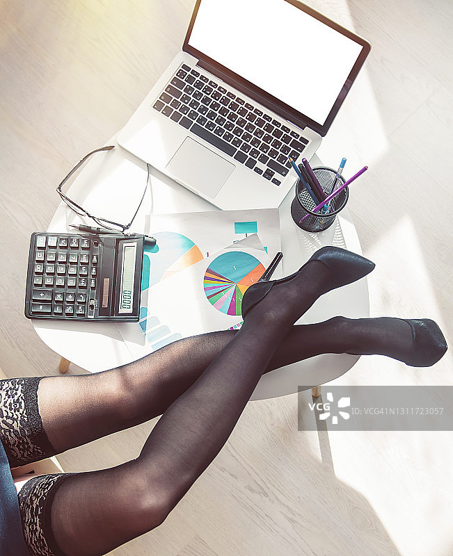 穿着黑色长筒袜和高跟鞋的女商人坐在沙发上，桌子旁放着一台笔记本电脑和财务图表。俯视图。图片素材