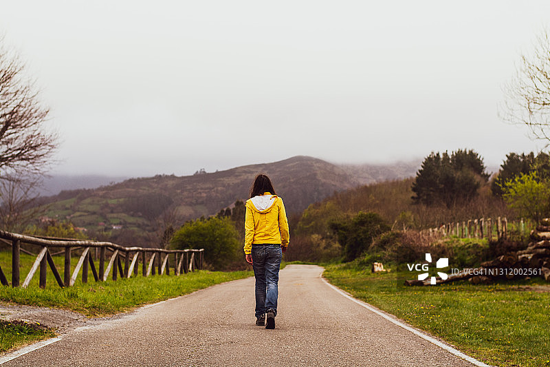 年轻女子身穿黄色雨衣和牛仔裤走在乡间小路中央的背影图片素材