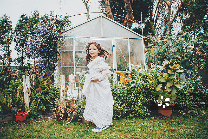 穿华丽连衣裙、头戴花冠的小女孩站在后院的温室旁图片素材
