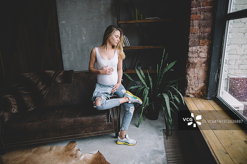 身穿白色泳衣和牛仔背带裤的金发年轻孕妇坐在阁楼式房间的皮沙发边缘，翘着二郎腿，双手捧着肚子图片素材