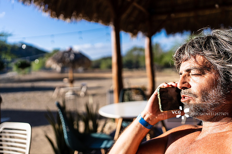 穿着泳衣的成年男子坐在海滩酒吧的椅子上用手机交谈图片素材