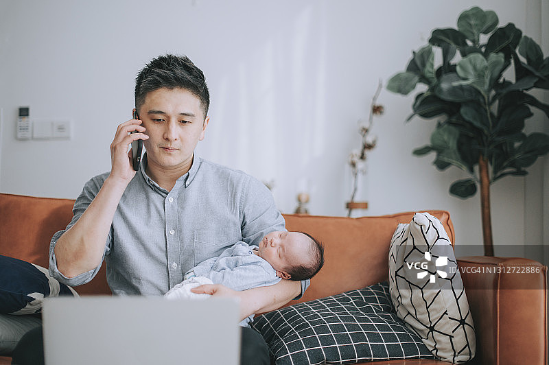 在家工作，在客厅沙发上抱着婴儿听电话的中国男人图片素材