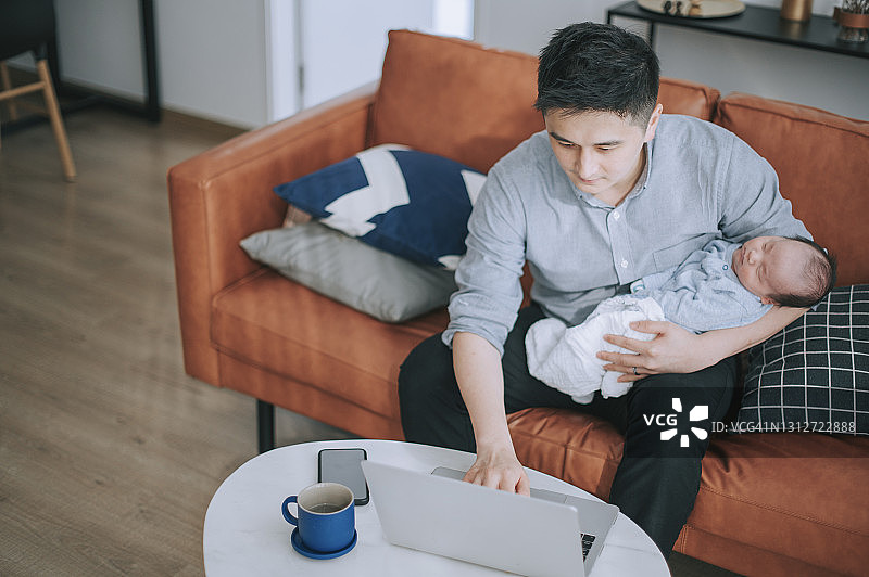 亚洲中国男人居家工作，在客厅沙发上抱着他的男婴使用笔记本电脑图片素材