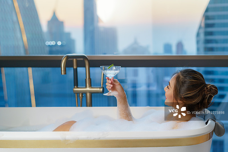 亚洲妇女在豪华酒店的浴缸里用肥皂泡泡洗澡，并在市中心度假时喝鸡尾酒图片素材