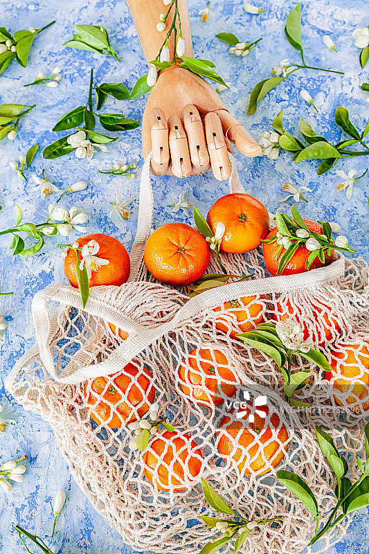 木手拿着装满橘子的棉网可重复使用购物袋，蓝色背景图片素材