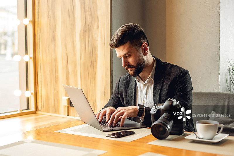 在咖啡馆使用笔记本电脑的成年白人男性，配有相机和饮品图片素材