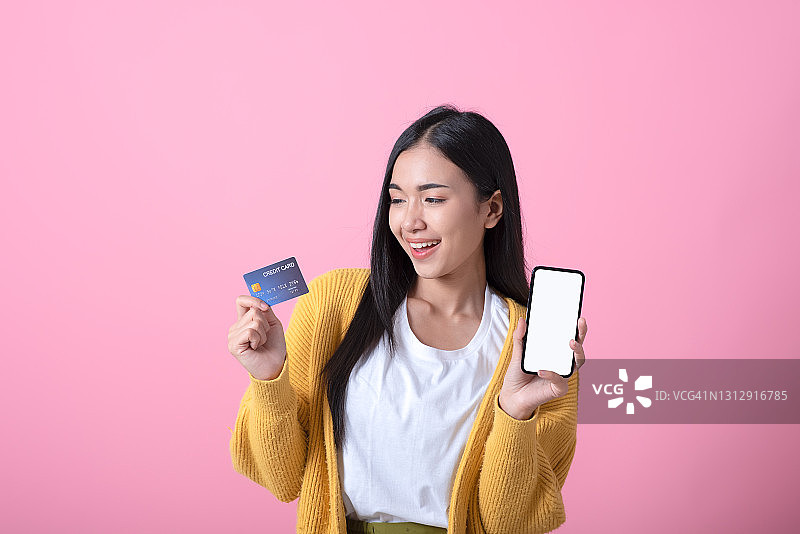 快乐的年轻女孩展示信用卡并手持手机（粉色背景）图片素材