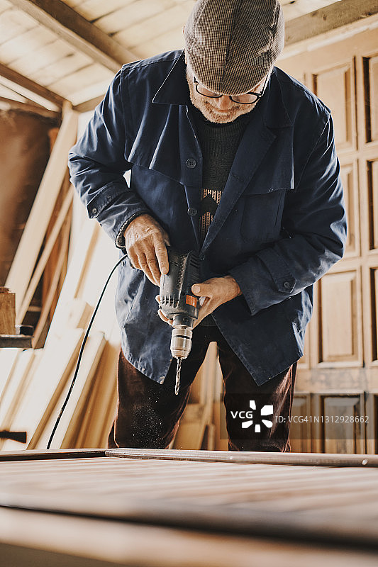 一位老木匠在旧车间里用钻头在木头上钻孔图片素材