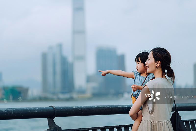 年轻漂亮的亚洲母亲抱着可爱的女儿，在海滨长廊欣赏壮观的城市天际线，享受亲子时光图片素材