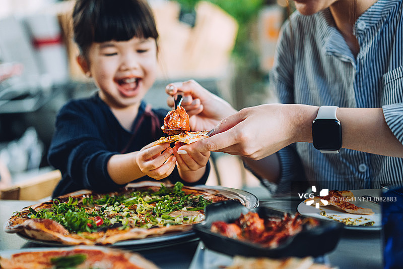 年轻的亚洲母亲和可爱的小女儿在户外餐厅享用披萨午餐，母亲为女儿提供肉丸和肉酱。家庭享受在一起的欢乐时光和美食。家庭和外出就餐的生活方式图片素材