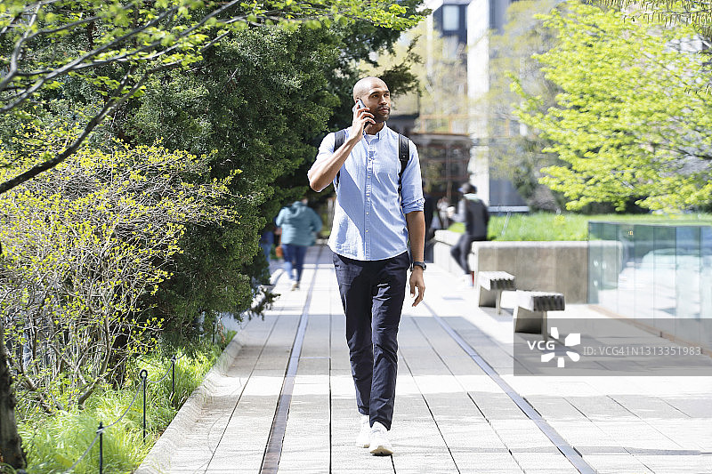 一位非裔美国千禧一代男人走在绿树成荫的地区，一边用手机通话，一边穿着商务休闲装，背着黑色背包。图片素材