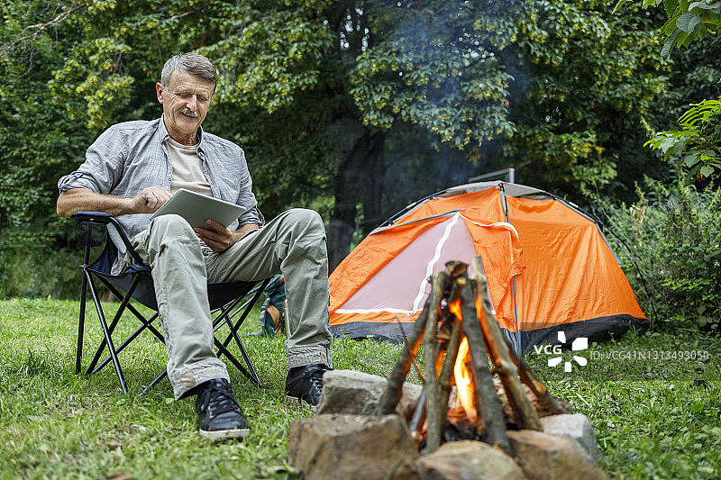 快乐的露营者坐在帐篷附近的篝火旁使用数码平板电脑图片素材