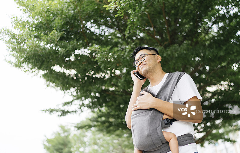 广角镜头下，年轻的亚洲父亲用手机在花园里拥抱背着婴儿背带的女儿图片素材