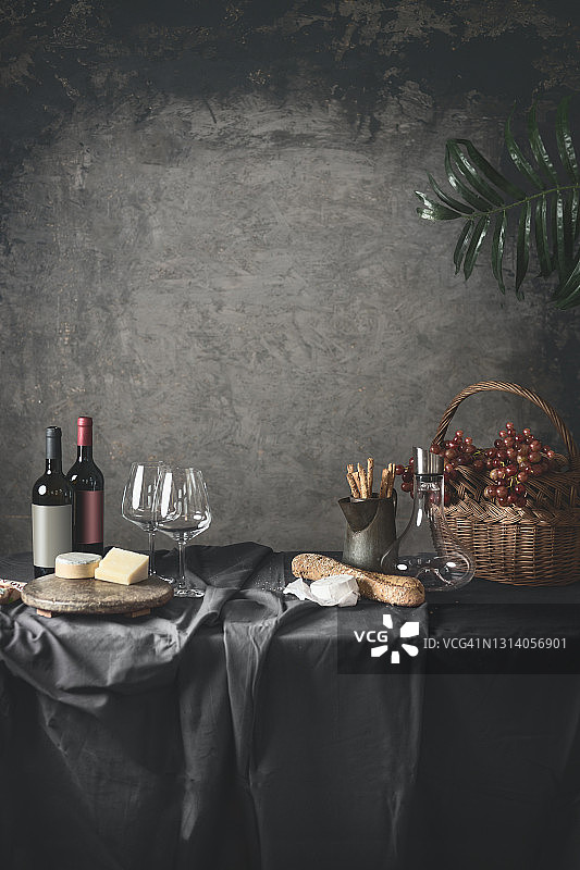 时尚静物，红酒瓶、奶酪、法棍面包、零食、葡萄在木篮里，葡萄酒杯和玻璃瓶放在灰色桌布的桌子上图片素材