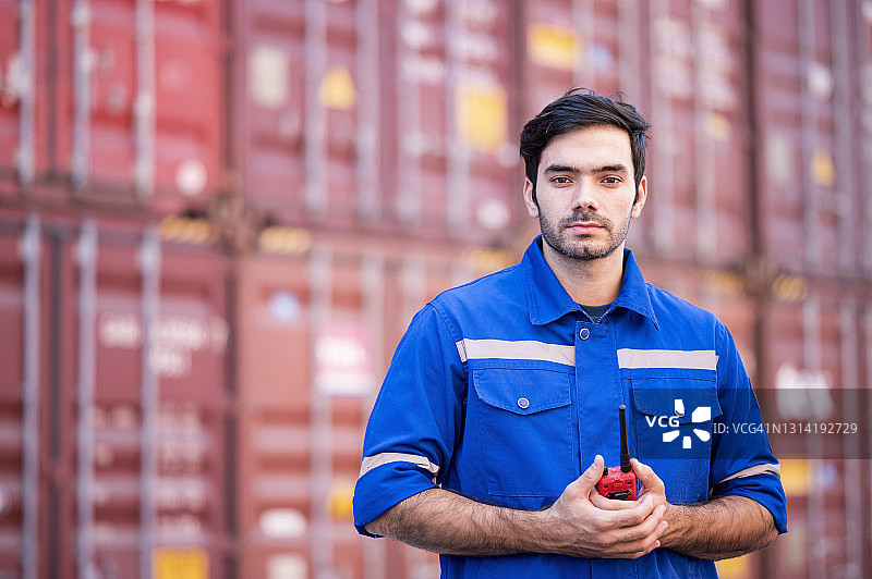 自信的工程师穿着工作服，手持对讲机，站在货运码头的集装箱堆垛前。进口、出口和货运代理业务概念图片素材