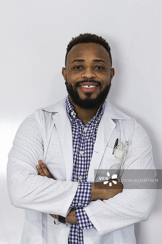 面带微笑的年轻男医生，双臂交叉站立于白墙前图片素材