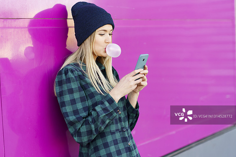 在墙前使用智能手机，吹着泡泡糖的格子连衣裙女人图片素材