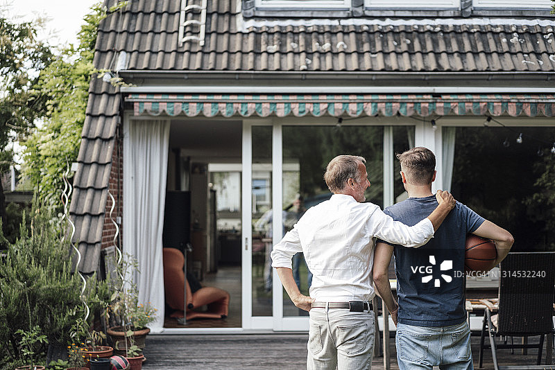 父亲搂着儿子，儿子抱着篮球站在后院图片素材