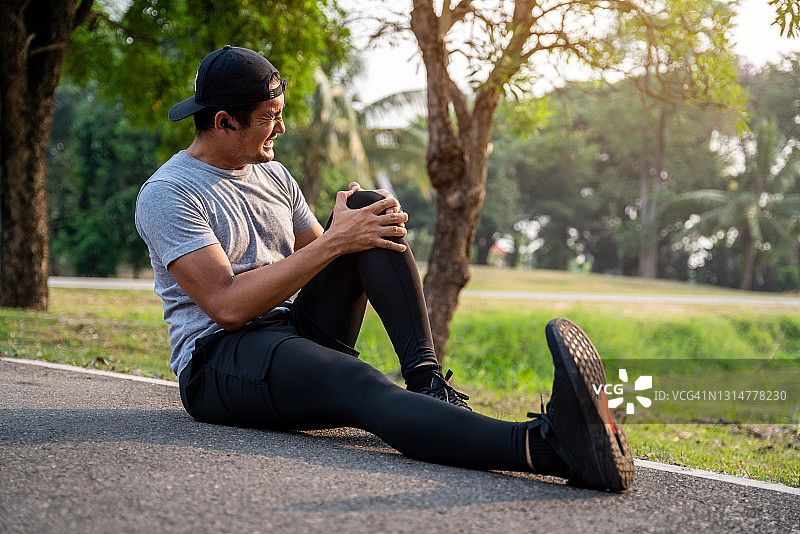 亚洲男性跑步者弯腰扶着膝盖，因在公园锻炼时膝盖急性受伤而感到剧烈疼痛，运动员膝盖受伤图片素材