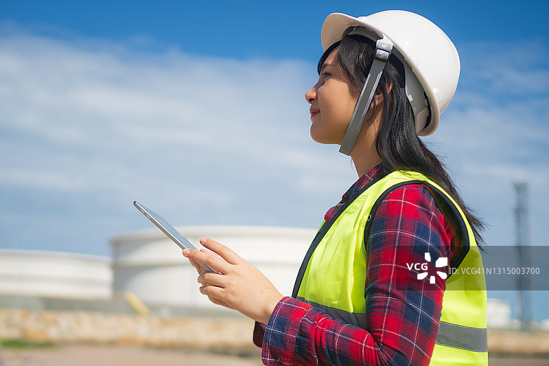 戴安全帽的女工程师在重油炼油厂油罐旁使用笔记本电脑图片素材