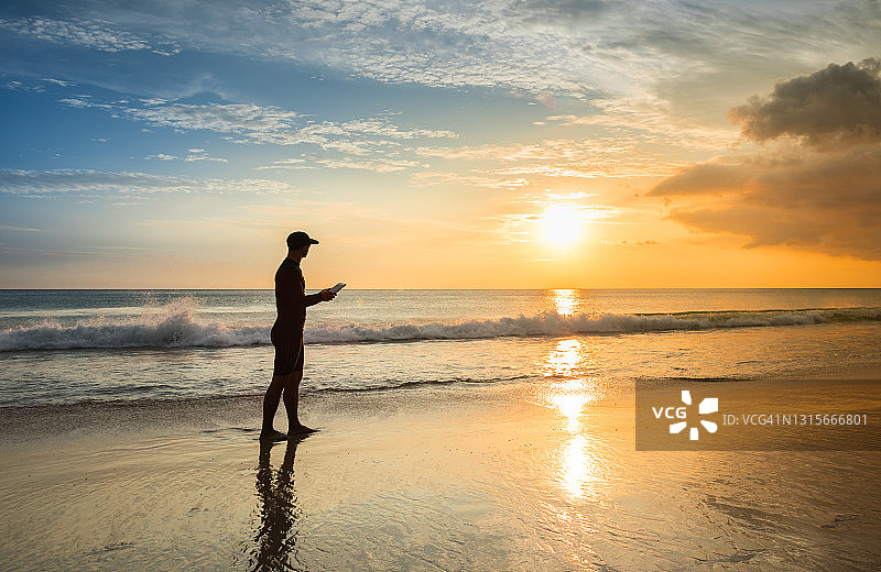 日落时分在海滩上使用平板电脑的年轻男人图片素材