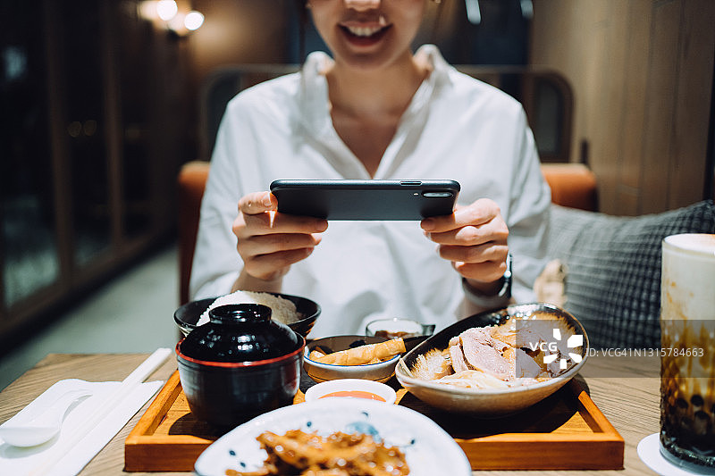 微笑的亚洲女人用智能手机拍摄美味的中国台湾风味套餐，包括各种开胃菜、卤鹅和珍珠奶茶，并在餐厅用餐前分享到社交媒体。外出就餐的生活方式。相机先吃文化图片素材