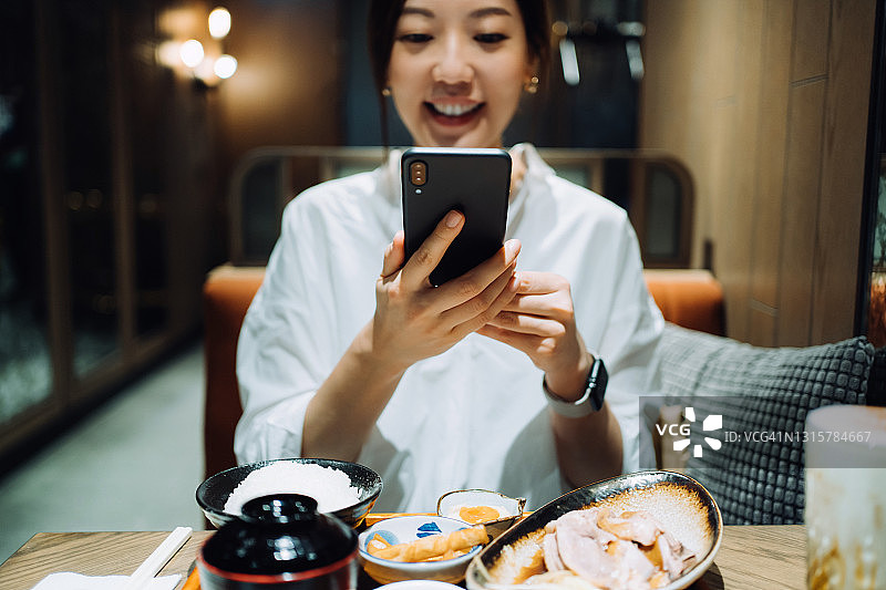 微笑的亚洲女人用智能手机拍摄美味的中国台湾风味套餐，包括各种开胃菜、卤鹅和珍珠奶茶，并在餐厅用餐前分享到社交媒体。外出就餐的生活方式。相机先吃文化图片素材