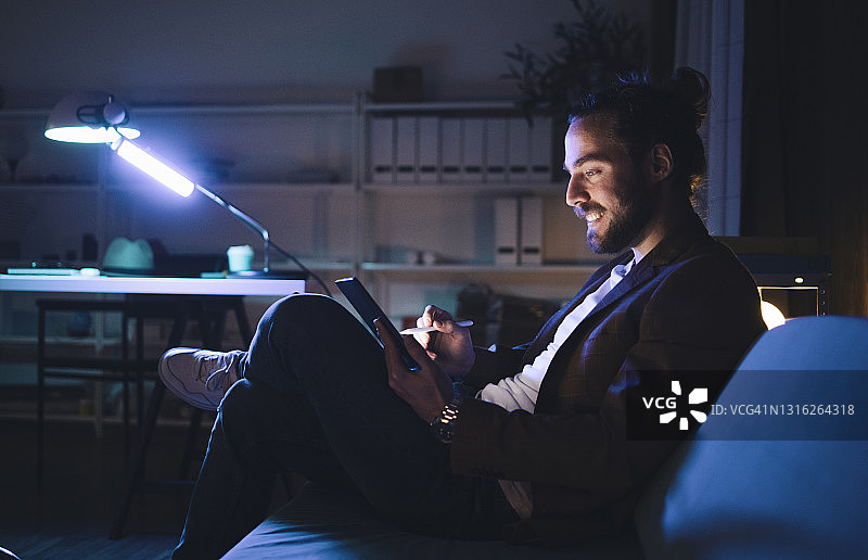 快乐的男人在黑暗中在沙发上使用平板电脑图片素材