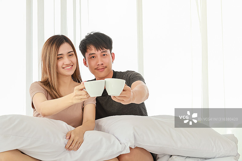 情人节概念：亚洲情侣端着热咖啡坐在卧室的床上图片素材
