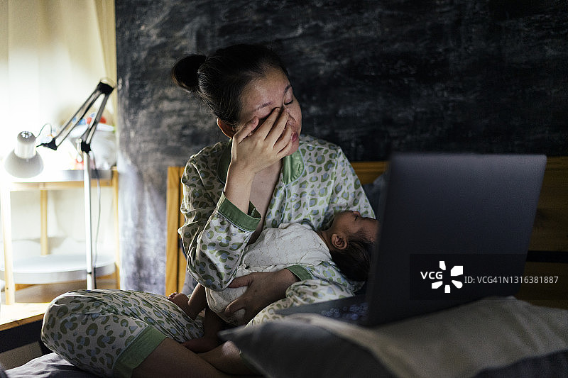 亚洲母亲深夜醒来给婴儿喂奶，并在床上点击笔记本电脑图片素材