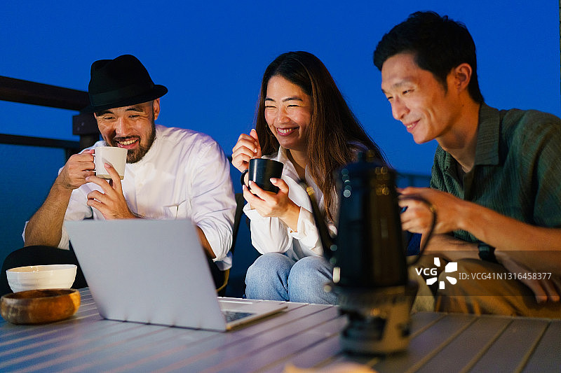 三个朋友在阳台夜晚喝热饮用笔记本电脑图片素材