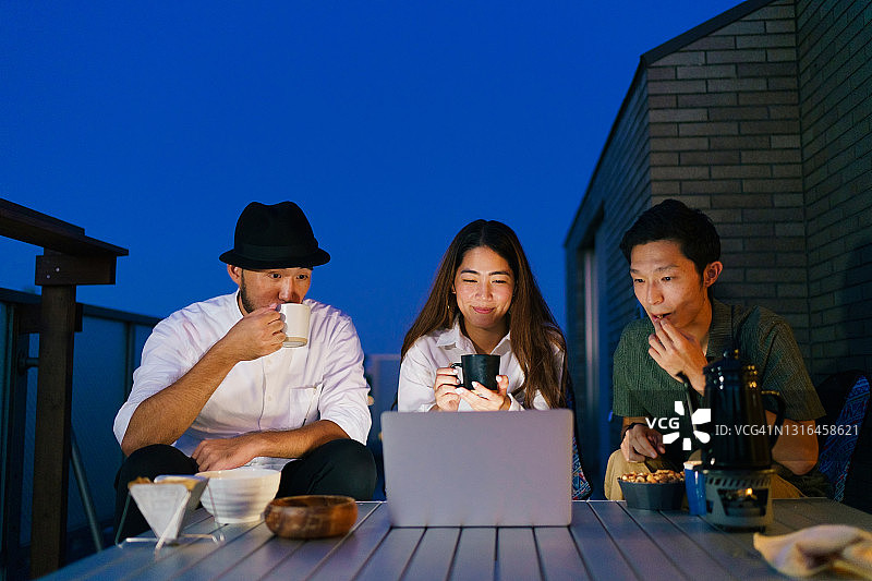 三个朋友在阳台夜晚喝热饮用笔记本电脑图片素材