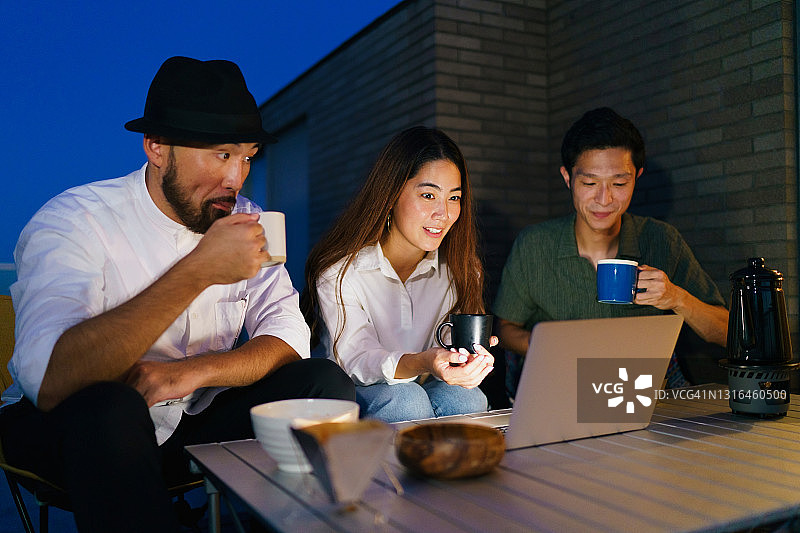 三个朋友在阳台夜晚喝热饮用笔记本电脑图片素材