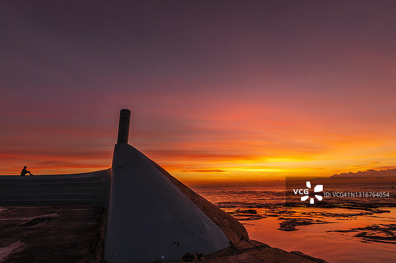 日出时海洋和海 pools 的美丽红色、黄色和橙色，远处有一个人坐在混凝土墙上图片素材