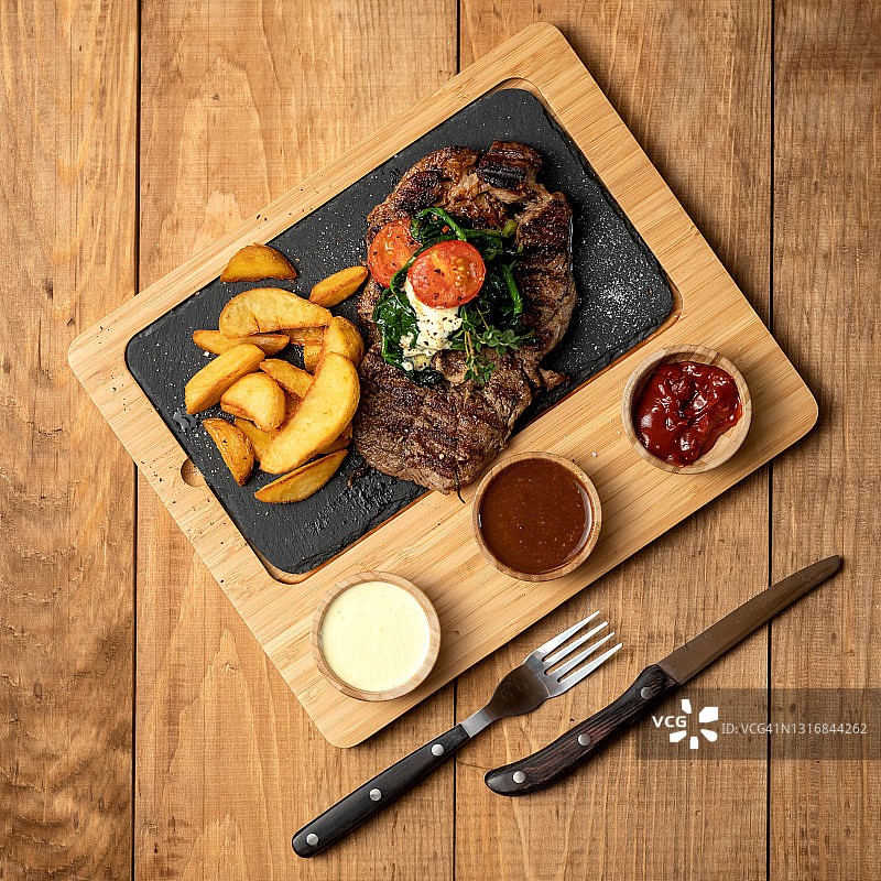配蔬菜的巨型多汁烤牛排。木制背景上的炸肉片配薯条和调味酱。顶视图图片素材