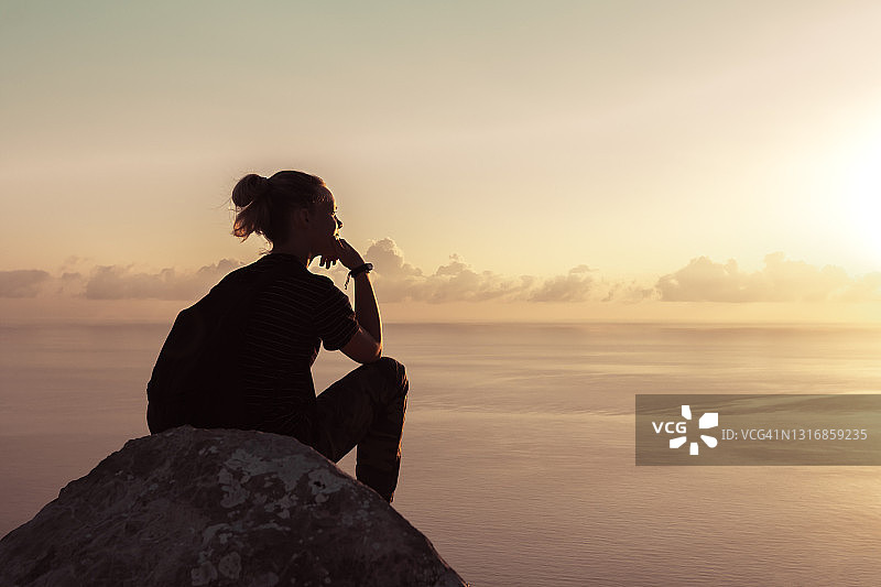 时髦女孩背着背包坐在海天背景的高高的岩石上图片素材