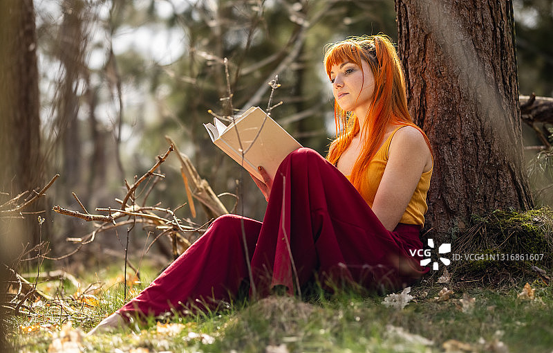 户外红发女孩在公园或花园里靠在树上看书图片素材