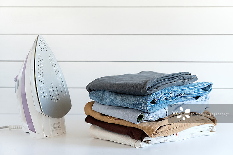 白色桌子或背景上的一堆物品，室内。干净的折叠棉质牛仔服装和彩色T恤。附近有一把电熨斗。家务、熨烫、护理和存放物品的概念。文本空间副本图片素材