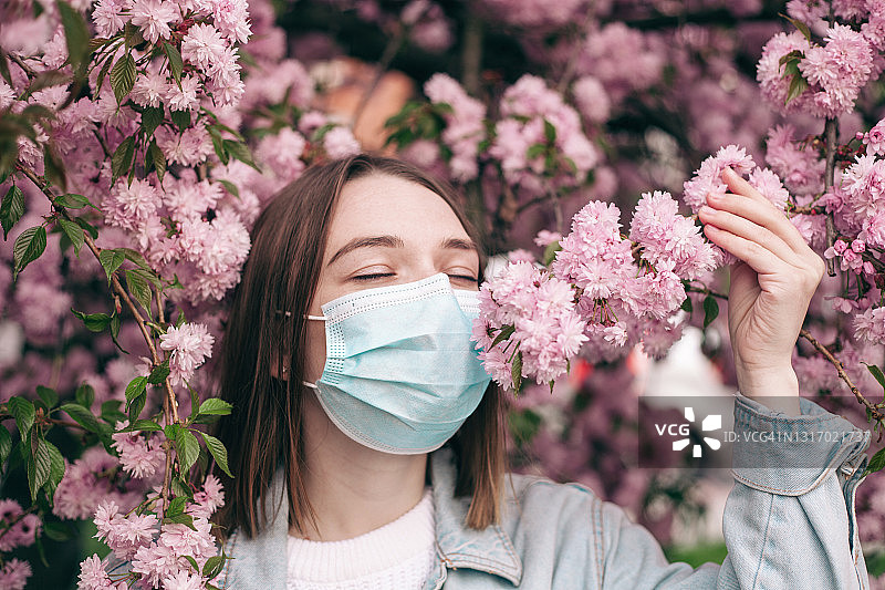戴着口罩嗅着粉色樱花的年轻女子，充满活力的粉色春天女性生活方式肖像图片素材