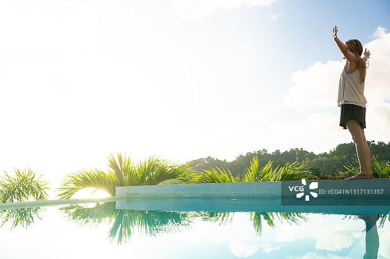 年轻女子站在丛林中豪华泳池边，举起双臂图片素材