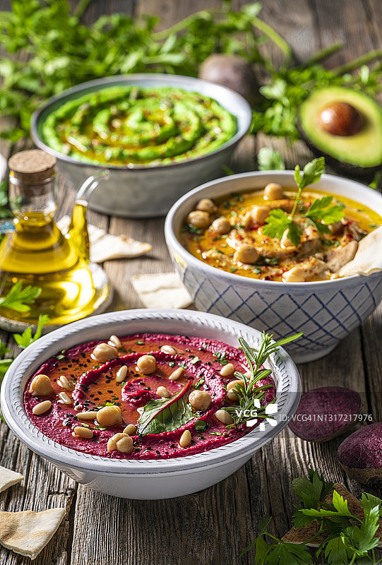 鹰嘴豆泥：三碗鹰嘴豆、鳄梨和甜菜根蔬菜，用于在乡村木桌上蘸酱图片素材