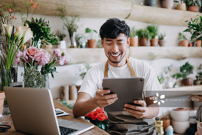 亚洲男花店老板使用平板电脑在笔记本电脑上工作，检查库存，接受客户订单，在线销售产品，利用科技经营小企业的日常图片素材