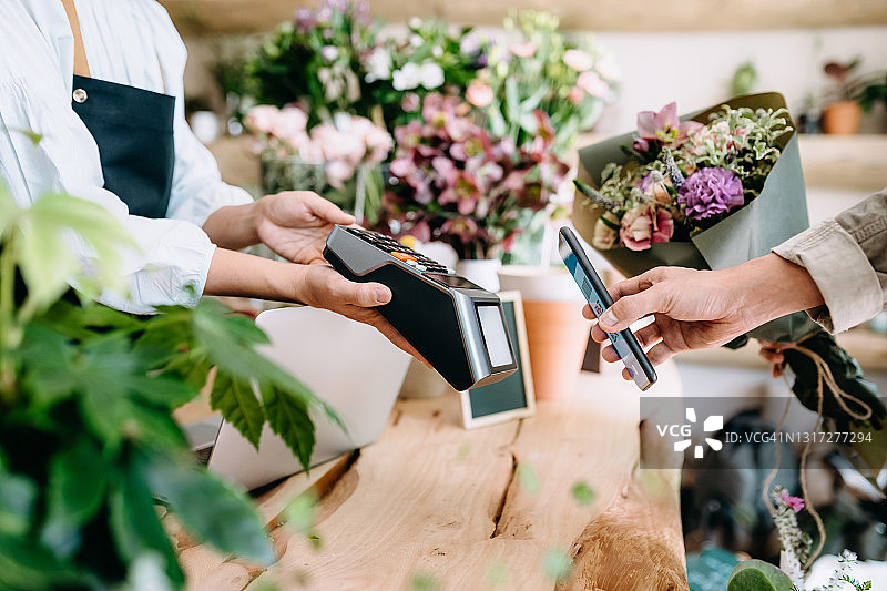 在花店购物的年轻亚洲男子特写。他正在用智能手机支付花束费用，在刷卡机上扫描并支付账单，实现快速简便的非接触式支付。NFC技术，即触即用概念图片素材