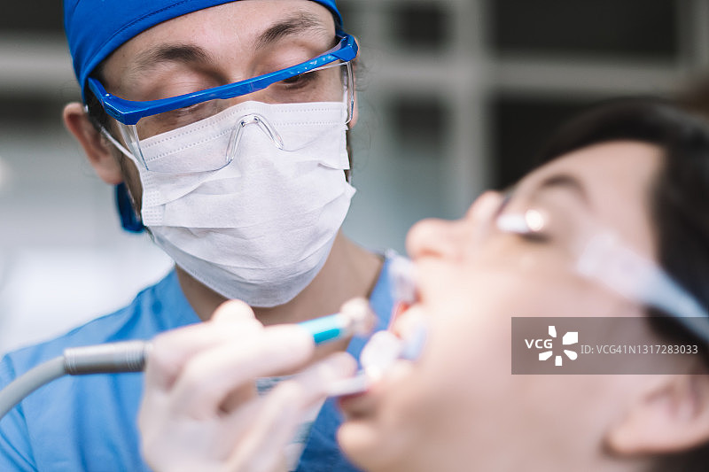 牙医正在治疗一位戴着防溅塑料眼镜和牙齿分离器的年轻女性的口腔图片素材