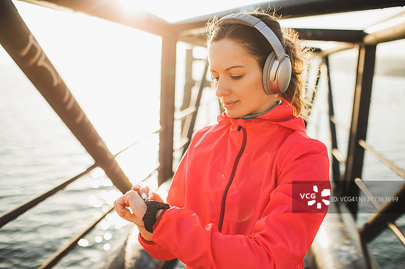 戴耳机的女人在智能手表或健身追踪器上启动跑步应用程序，城市风格，日出图片素材