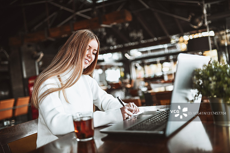 微笑的金发女性在餐厅喝茶时用笔记本电脑学习图片素材