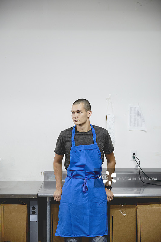 在工厂靠墙站立，身穿蓝色围裙的沉思工人图片素材