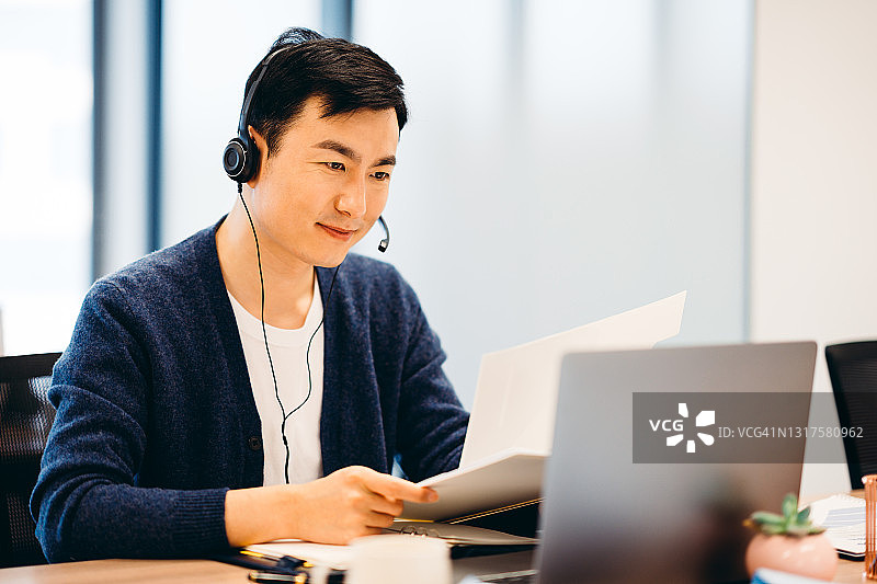 亚洲商人戴着耳机在办公室里参加视频会议。图片素材