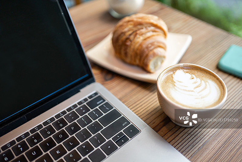 咖啡馆桌上的拉花咖啡、笔记本电脑和羊角面包图片素材