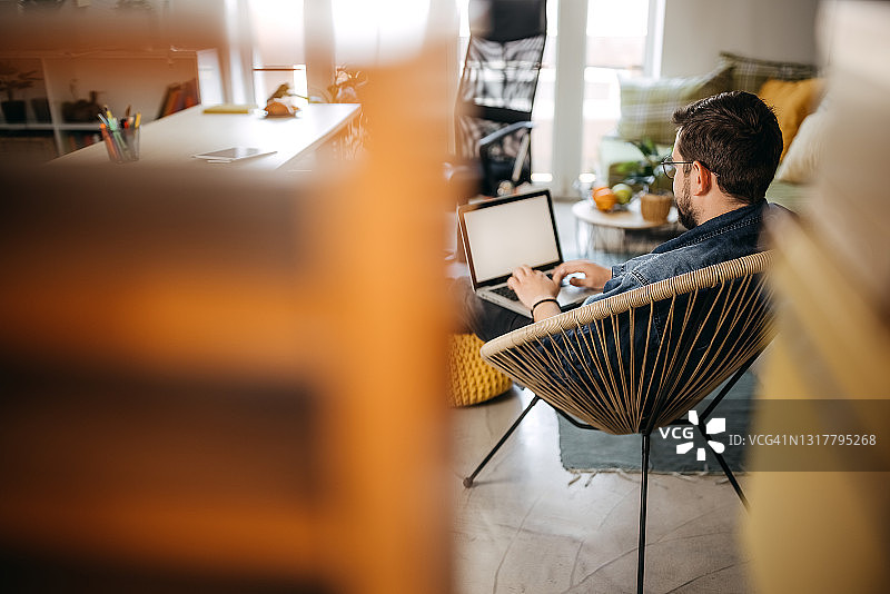 戴眼镜的年轻络腮胡男子坐在家中的扶手椅上使用笔记本电脑图片素材